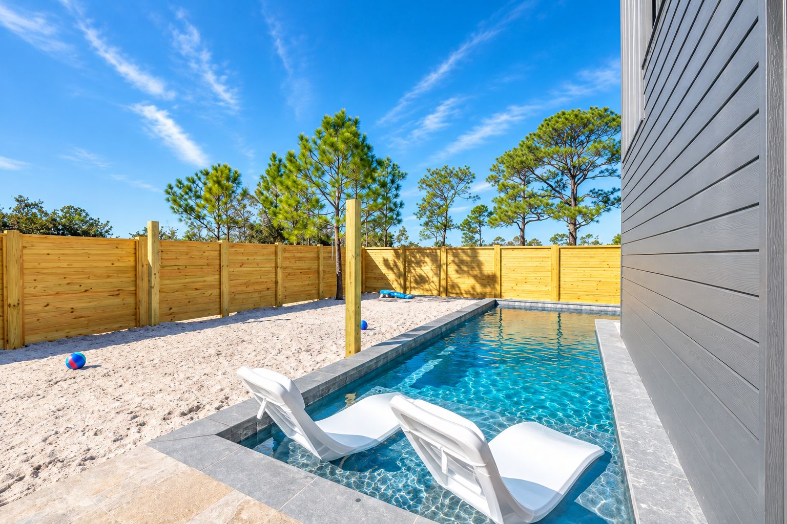A swimming pool with two lounge chairs, a wooden fence, and trees in the background.