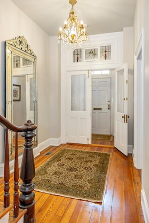 A hallway with wooden flooring, a large ornate mirror, a chandelier, and a patterned rug.