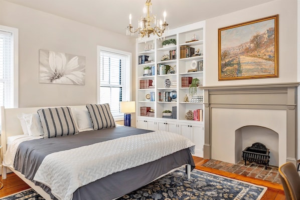 A bedroom with a large bed, a bookshelf, a fireplace, and a painting on the wall.