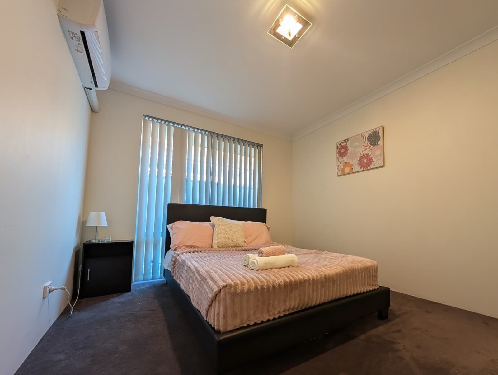 A bedroom with a bed, a nightstand, an air conditioner, a window with blinds, and a wall-mounted light fixture.