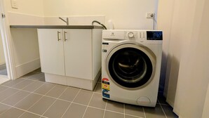 Kompakti pyykinhuoltotila, jossa on etuaukosta täytettävä pesukone, pieni kaappi ja laatoitettu lattia.