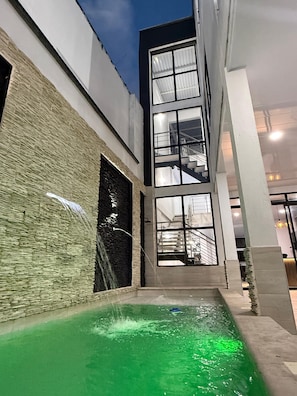Un edificio moderno con fachada de vidrio, una pequeña piscina al aire libre con agua verde y un muro de piedra en un lateral.