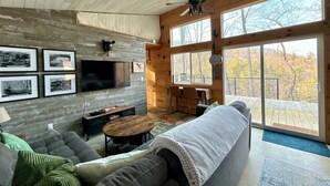 A cozy living room with a large TV, wooden walls, a ceiling fan, and a view of the outdoors.