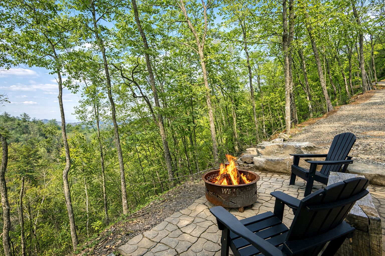 A fire pit with two Adirondack chairs, surrounded by a stone patio and dense forest.
