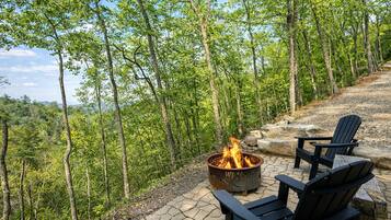 A fire pit with two Adirondack chairs, surrounded by a stone patio and dense forest.