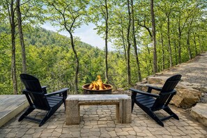 An outdoor seating area with two Adirondack chairs, a stone fire pit, and a wooden table. - Wrigs Ridge | Tiny Cabin w/ Views & Creek Sound (Robbinsville)