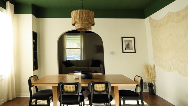 Un comedor con mesa de madera, cuatro sillas, lámpara de fibra, alfombra a cuadros y un tapiz grande en la pared.
