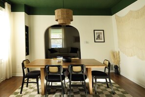 Un comedor con mesa de madera, cuatro sillas, lámpara de fibra, alfombra a cuadros y un tapiz grande en la pared.
