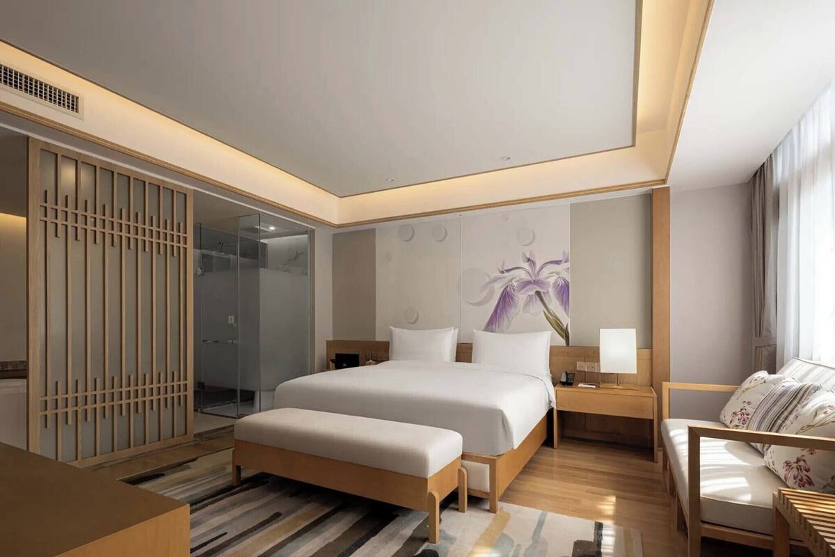 A modern hotel room with a large bed, bedside tables, a seating area, and a glass shower enclosure.