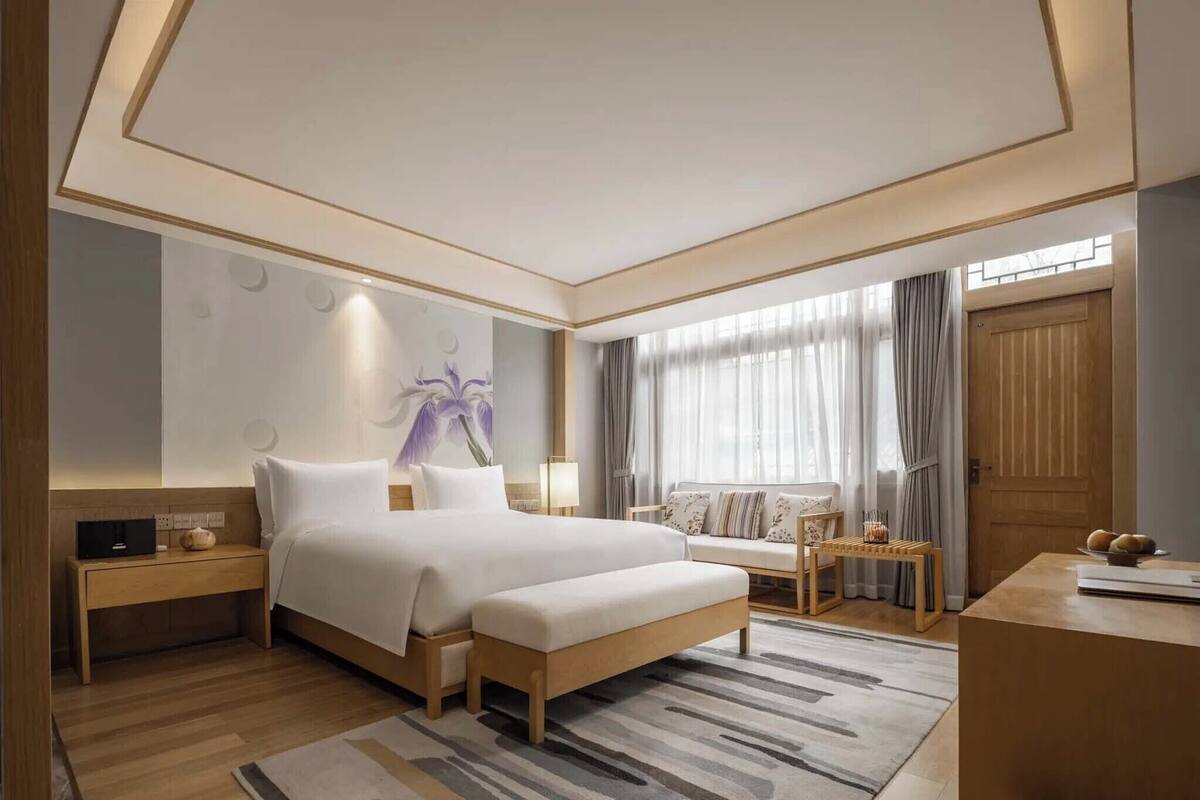 A modern hotel room with a large bed, bedside tables, a seating area with a sofa, and a wooden door.