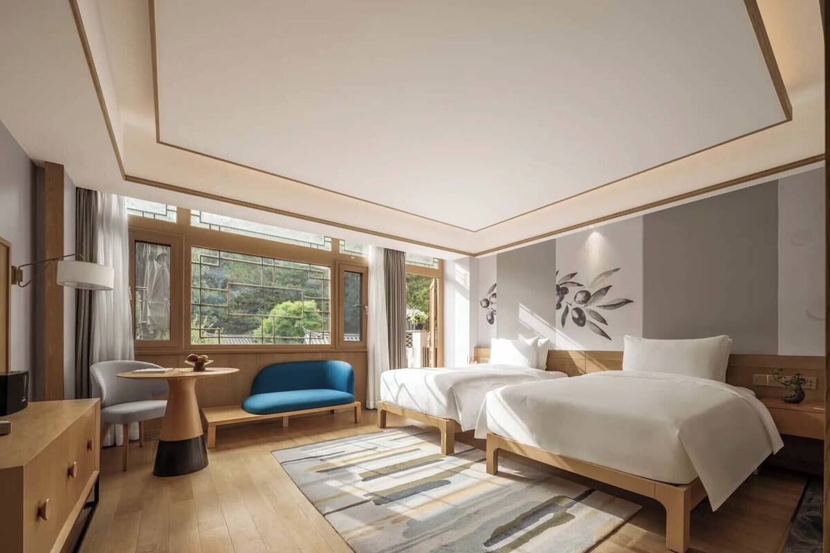 A modern hotel room with a large bed, a sofa, a desk, and a window with a view of greenery.