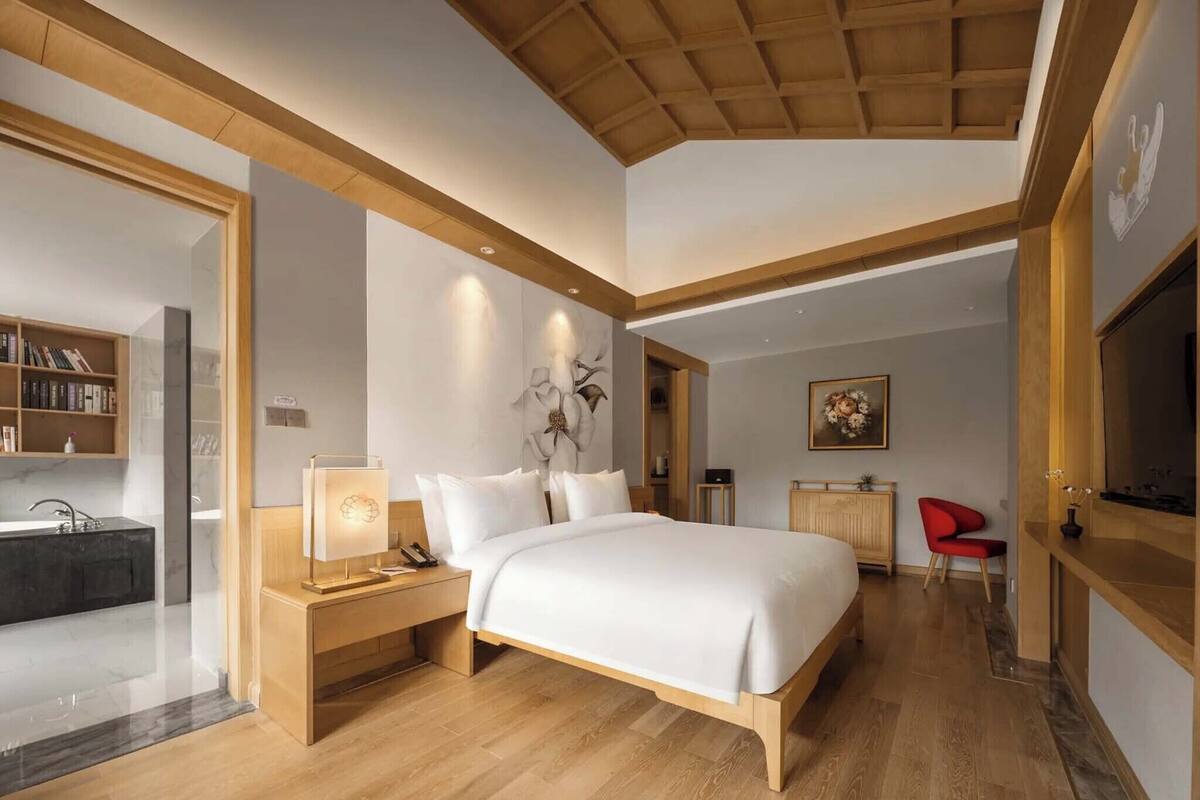 A modern bedroom with a wooden ceiling, a large bed, a bedside table with a lamp, a red chair, and a painting on the wall.