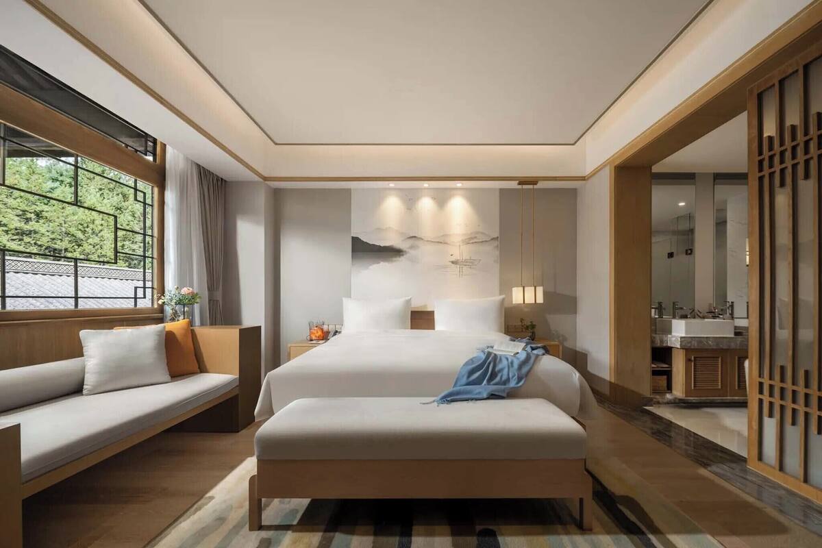 A modern hotel room with a large bed, a sofa, a bathroom with a sink, and a view of trees through a window.
