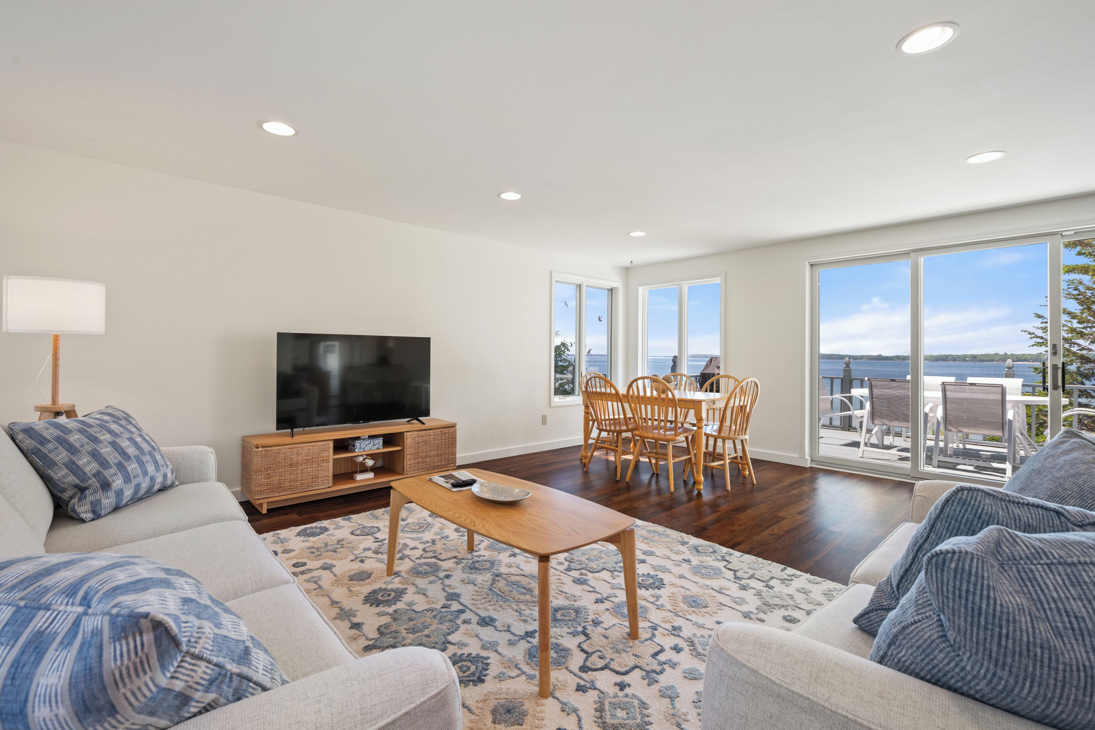 Amplia sala de estar con televisor de pantalla plana, comedor con sillas de madera y ventanas grandes con vista al mar.