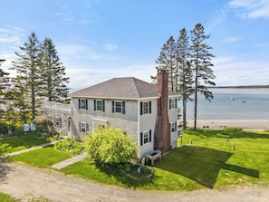 A two-story house with a large chimney, a grassy yard, and a view of the water. - Private Oceanfront Oasis 4-bedroom house in Brooklin perfect for your getaway (Brooklin)