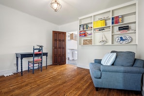 A room with a wooden floor, a blue armchair, a desk with a chair, and a bookshelf filled with books and games. - Private Oceanfront Oasis 4-bedroom house in Brooklin perfect for your getaway (Brooklin)