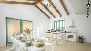 A dining area with a white table set for a meal, white chairs, a chandelier, and a kitchen in the background. - Villa Kaia (Istria - Kringa) (Kringa)