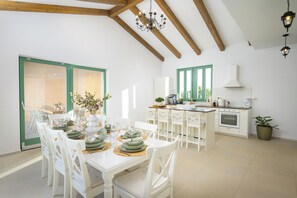 Ein Essbereich mit einem weißen Tisch, gedeckt für eine Mahlzeit, weißen Stühlen, einer Chandelierleuchtre und einer Küche im Hintergrund.
