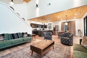 Ein geräumiger Wohnbereich mit einem grünen Sofa, gemusterten Sesseln, einem Holztisch und einer Küche im Hintergrund.