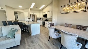 Une cuisine moderne avec des meubles blancs, un réfrigérateur en acier inoxydable et un coin repas comprenant une table en bois clair et des chaises à revêtement gris.