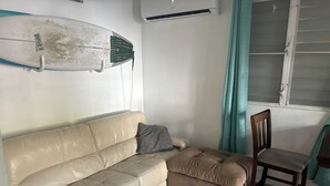 Un canapé en cuir beige avec un coussin, une chaise en bois, une planche de surf accrochée au mur et une fenêtre avec des rideaux.