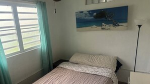 Une chambre à coucher avec un lit, une fenêtre avec des rideaux, un tableau représentant une scène de plage et un ventilateur de plafond.