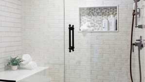 Et moderne badeværelse med et brusebadekar i glas, hvide flisemure og en lille hylde med toiletartikler.