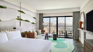 Habitación de hotel con una cama grande, un sofá, una mesita, un televisor y vistas a las montañas.