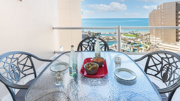 Ein Tisch mit Glasplatte, ein roter Tablett, eine Flasche und eine Oliven-Schale auf einem Balkon mit Blick auf Strand und Resort.
