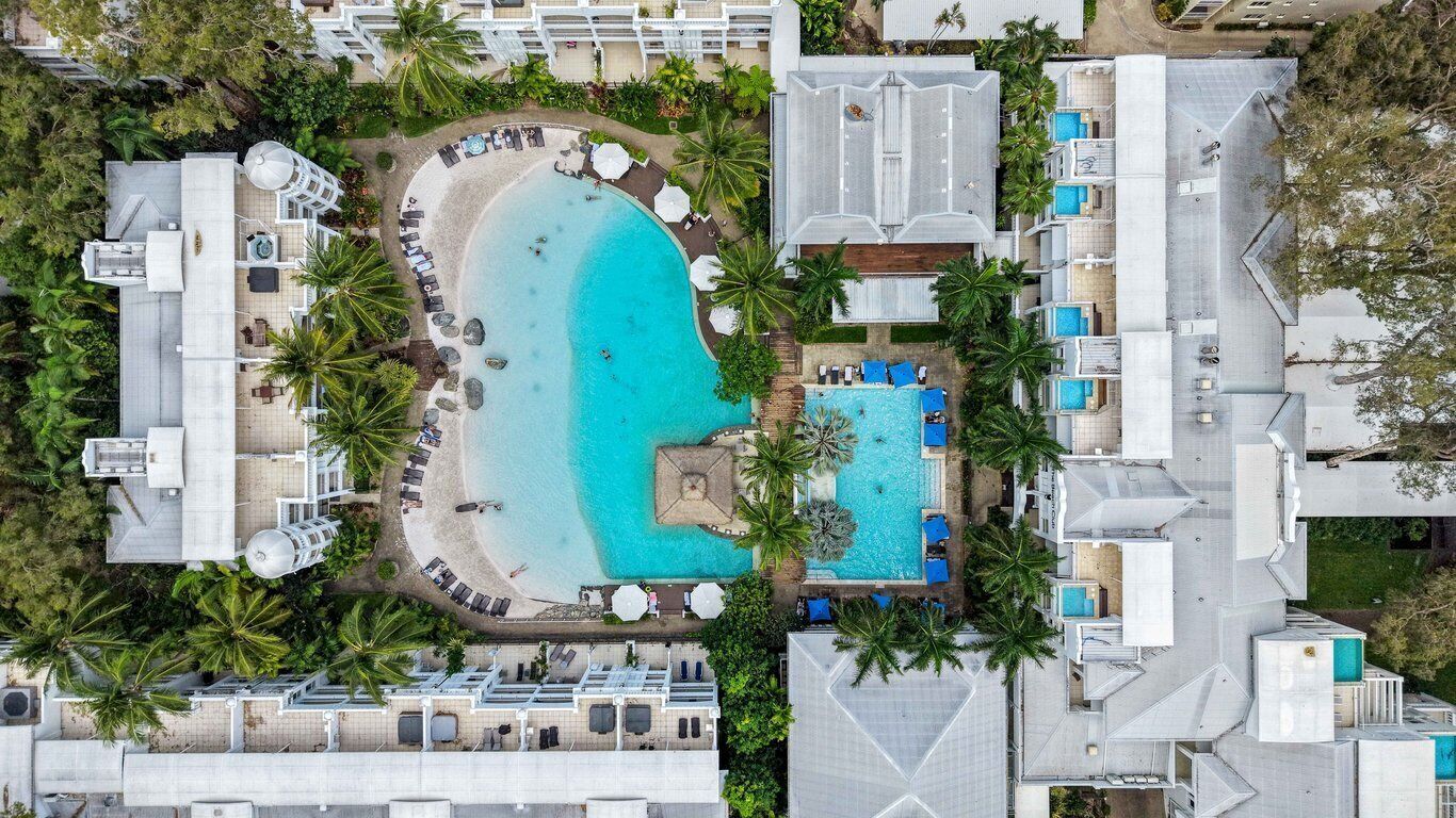 Luchtbeeld van een resort met een groot centraal zwembad, meerdere gastenkamers en weelderige tropische beplanting.