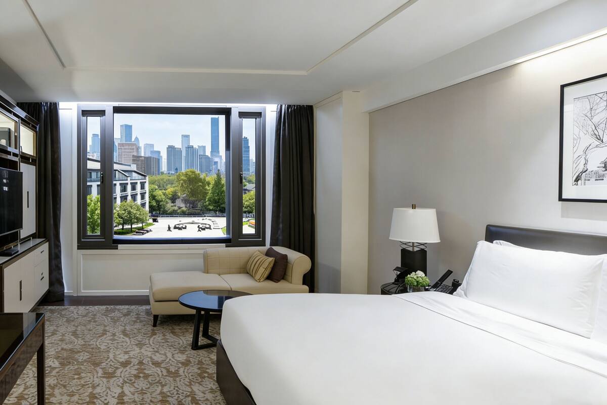 Hotelkamer met een bed, een bank, een kleine tafel, een bureau met lamp, een televisie en een raam met uitzicht op de stad.