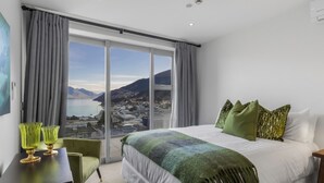 Ein Schlafzimmer mit einem großen Bett, grünen Kissen und Blick auf eine Berglandschaft. Ein Tisch mit grünem Glas steht ebenfalls im Raum.