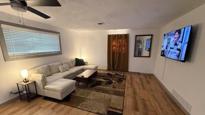 Ein Wohnzimmer mit einem Ecksofa, einem Couchtisch, einem Teppich, einem Deckenventilator, einem Fernseher und einem Fenster mit Jalousien.