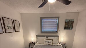 Ein Schlafzimmer mit einem Bett, zwei Nachttischen, einem Deckenventilator, einem Fenster mit Jalousien, gerahmten Pflanzenbildern und einem Wandbild.