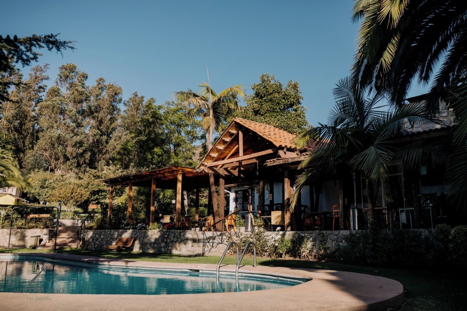 Area piscina con una pergola in legno, un muro in pietra e un edificio con tetto in tegole.
