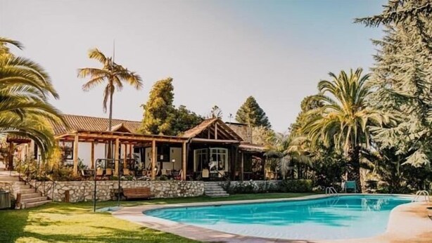 Un espace aménagé au bord de la piscine, avec une maison, un mur en pierre et des palmiers.
