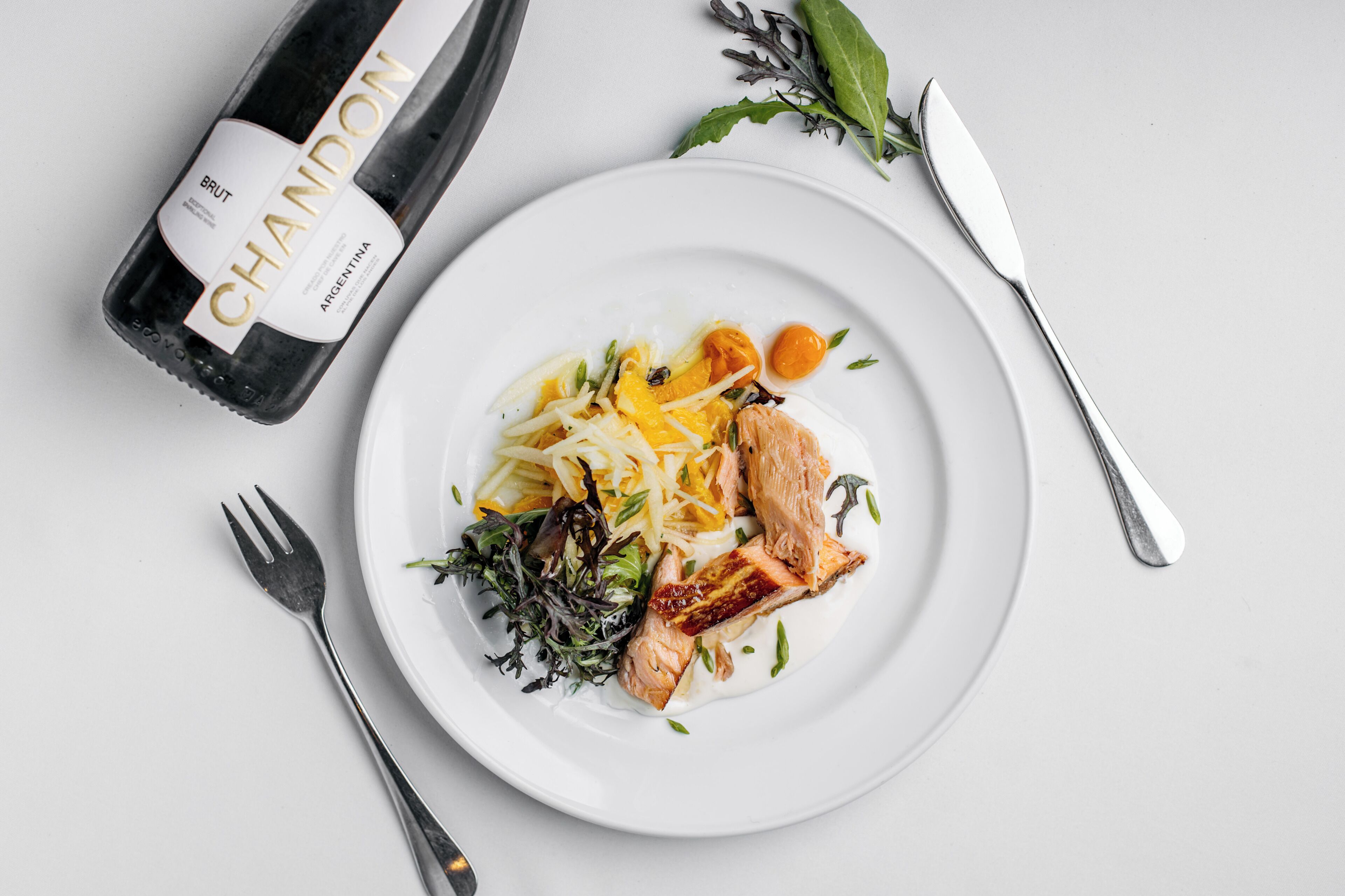 Ein Teller mit Essen, eine Flasche Chandon Brut Wein, eine Gabel und ein Messer auf weißer Oberfläche.