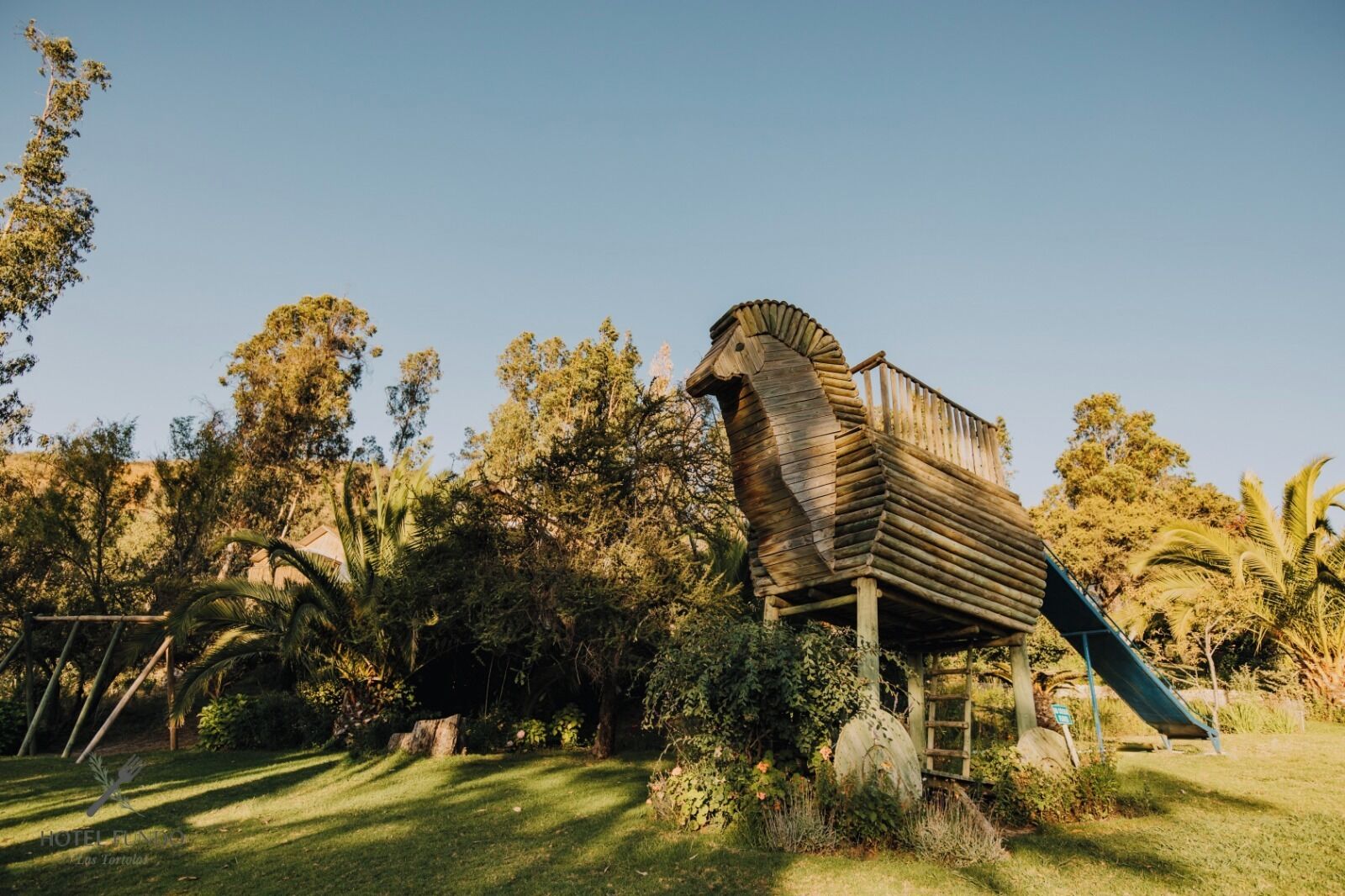 A wooden horse sculpture with a slide, surrounded by lush greenery and trees.