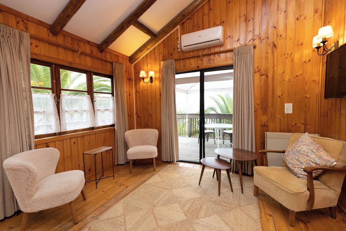 A wooden-paneled room with two armchairs, a sofa, a small table, and a window with curtains.