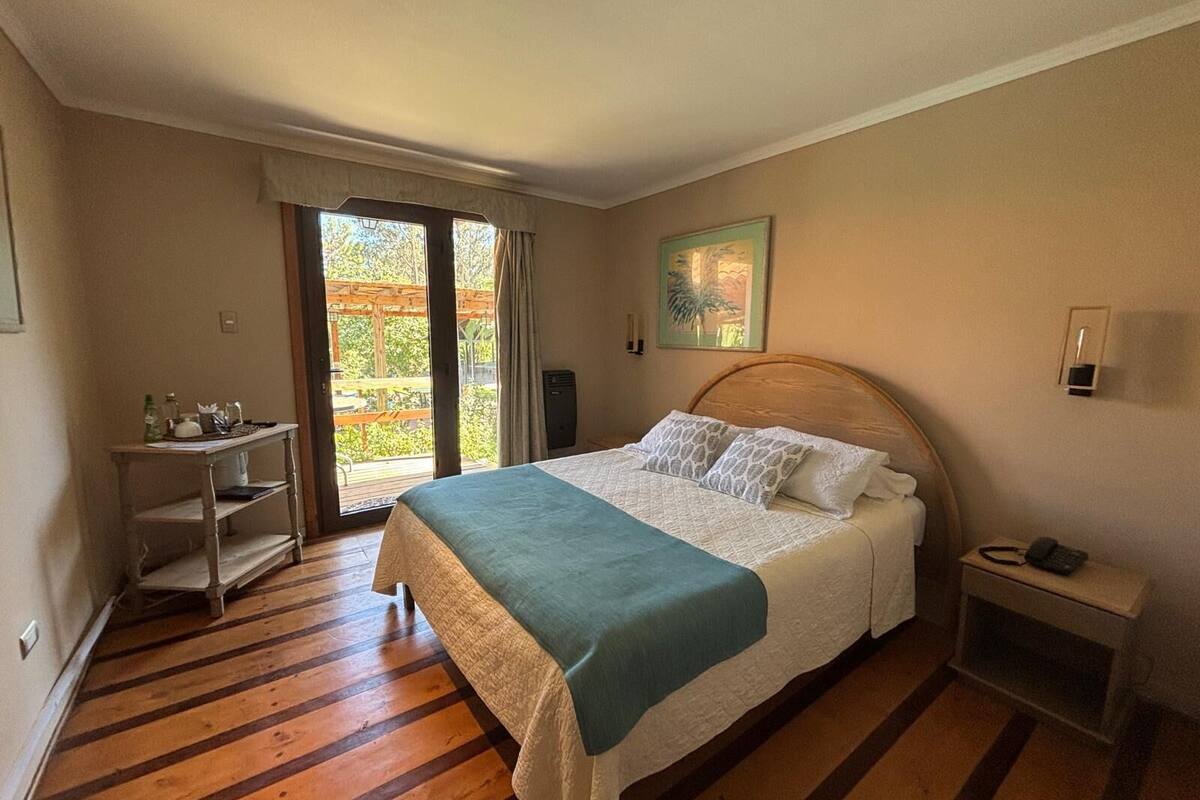 A bedroom with a bed, a bedside table, a nightstand, a window with a view, and a door leading to an outdoor area.