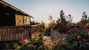 Un bâtiment rustique en bois, doté d’un balcon, entouré d’un jardin où poussent des fleurs roses et bordé d’un mur de pierres.