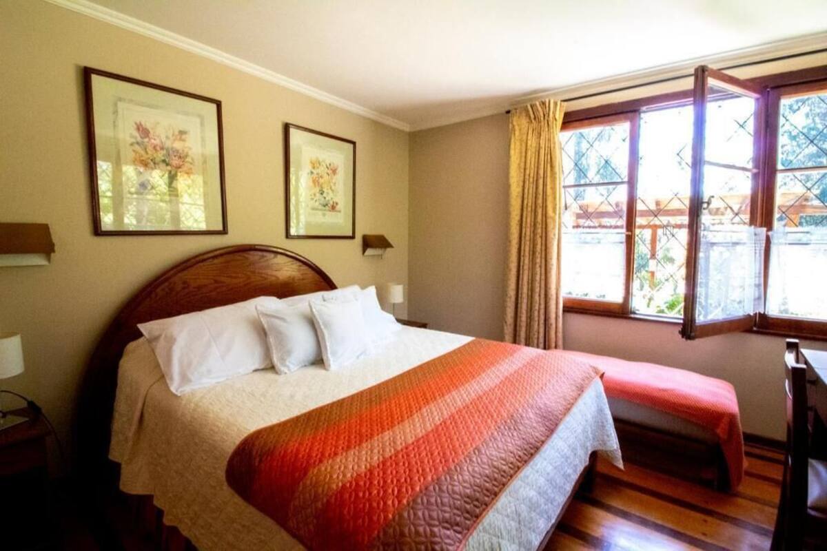 A hotel room with a large bed, a bench, a window with curtains, and two framed pictures on the wall.