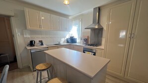 Ett modernt kök med vita skåp, en köksö, två barstolar, en diskmaskin, en spis och ett fönster med gardiner.