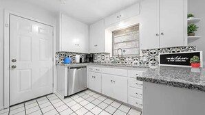 Una cocina moderna con armarios blancos, lavavajillas de acero inoxidable, salpicadero con diseño blanco y negro, y un cartel que dice 'Esta cocina es para bailar'.