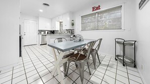 Cocina moderna con armarios blancos, una mesa de comedor con sillas, una ventana con persianas y un cartel que dice 'Eat'.