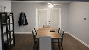 En spisestue med et træbord, seks stole, en loftsvifte og en reol med bøger og jakker.