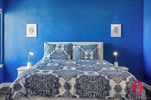 Une chambre à coucher avec une couvre-lit à motifs bleus et blancs, deux lampes de chevet et des œuvres d’art encadrées accrochées au mur.