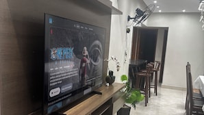 A modern living room with a mounted TV, wooden entertainment unit, and a dining area with chairs and a table. - Beautiful and cozy duplex with private pool. (Guarujá)