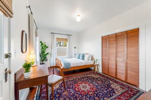 A bedroom with a bed, wooden wardrobe, a desk with a plant, a chair, a window with curtains, and a patterned rug.