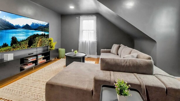 A modern living room with a large flat-screen TV, a grey sofa, a small round table, and a bookshelf.